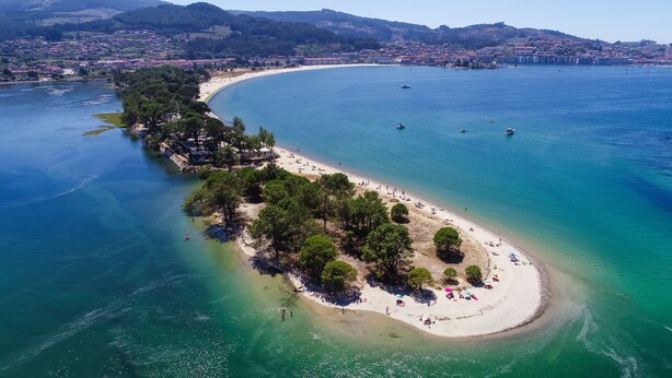 Vista aérea de una zona costera con playa arenosa, aguas azul cristalino y una pequeña isla con árboles.