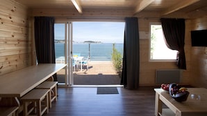 Una cabaña de madera con un amplio ventanal corredera que da a un patio con vistas al mar. En el interior, hay una mesa de madera y un banco.