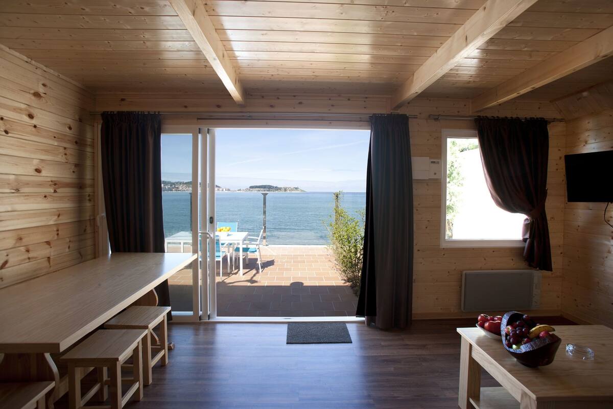 Eine Holzhütte mit einer großen Schiebetür, die auf eine Terrasse mit Meerblick führt. Im Inneren befindet sich ein Holztisch und eine Bank.