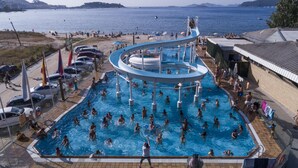 Una gran piscina al aire libre con un tobogán acuático, rodeada de gente disfrutando en la playa y coches estacionados.