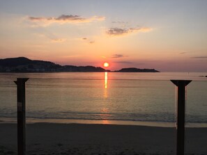 A sunset over a calm sea with a beach in the foreground and a distant shoreline with buildings. - Camping Bayona Playa (Baiona)
