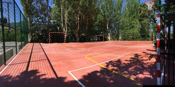 An outdoor basketball court with a red surface, surrounded by trees and a green fence.