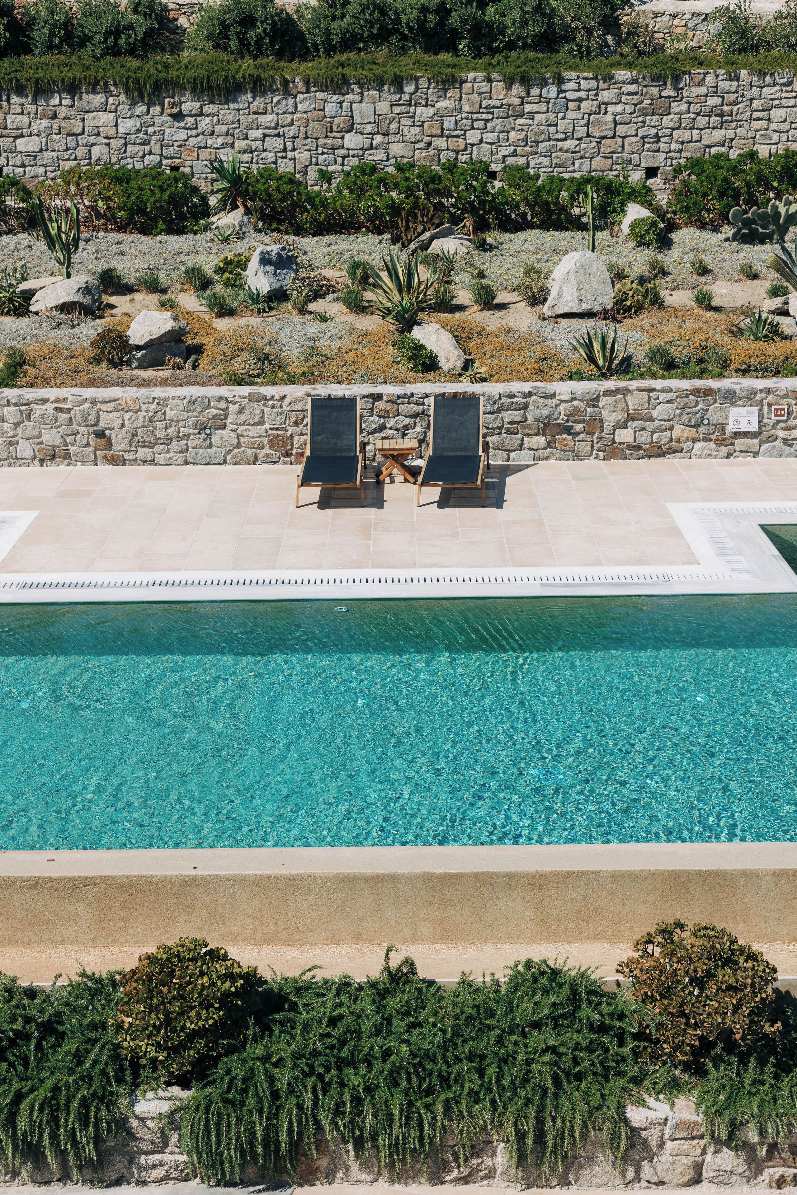 A pool with two lounge chairs, a stone wall, and a variety of plants.