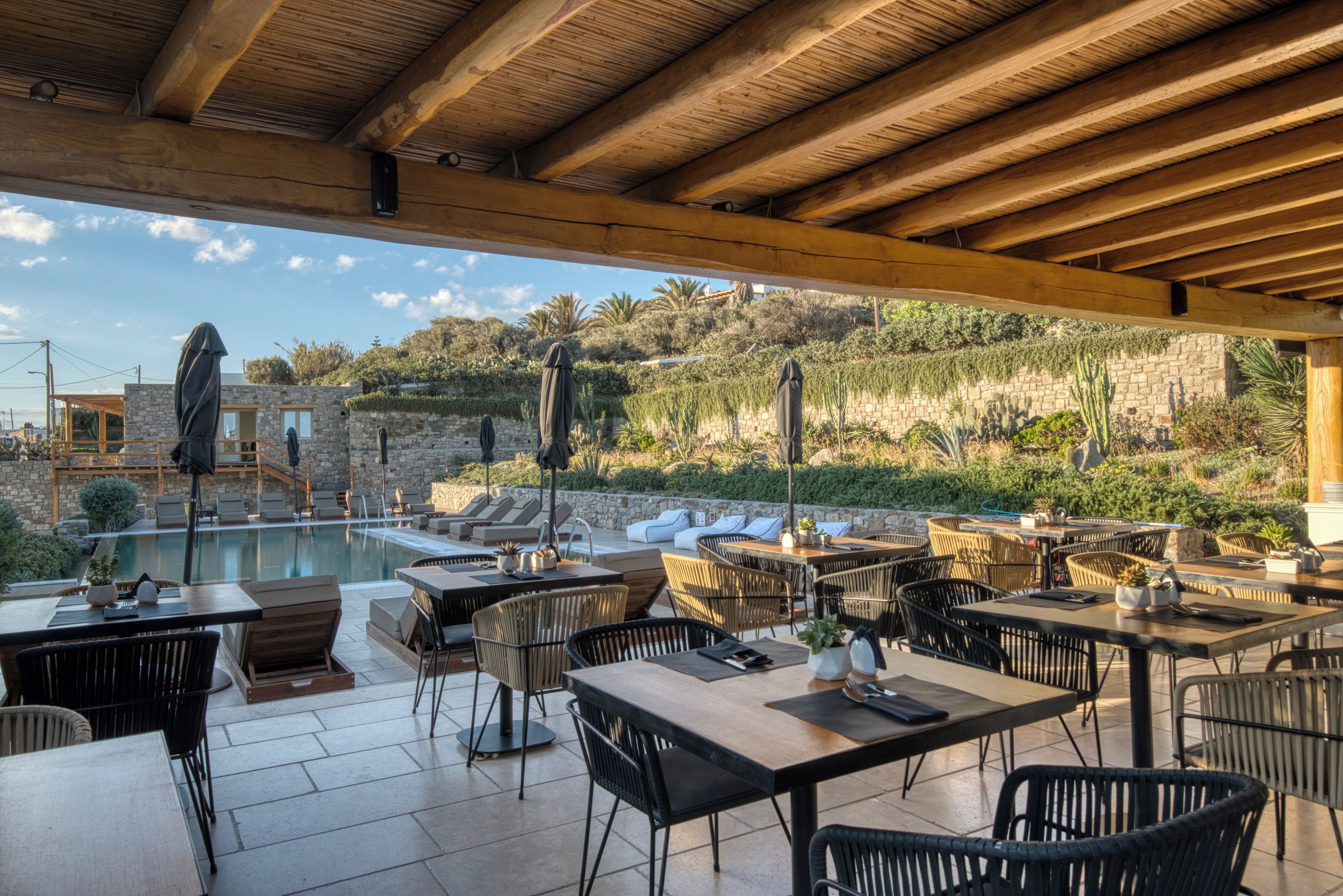 Un espace de restauration en plein air, avec des tables et des chaises, donnant sur une piscine et un jardin aménagé avec des cactus et des arbustes.