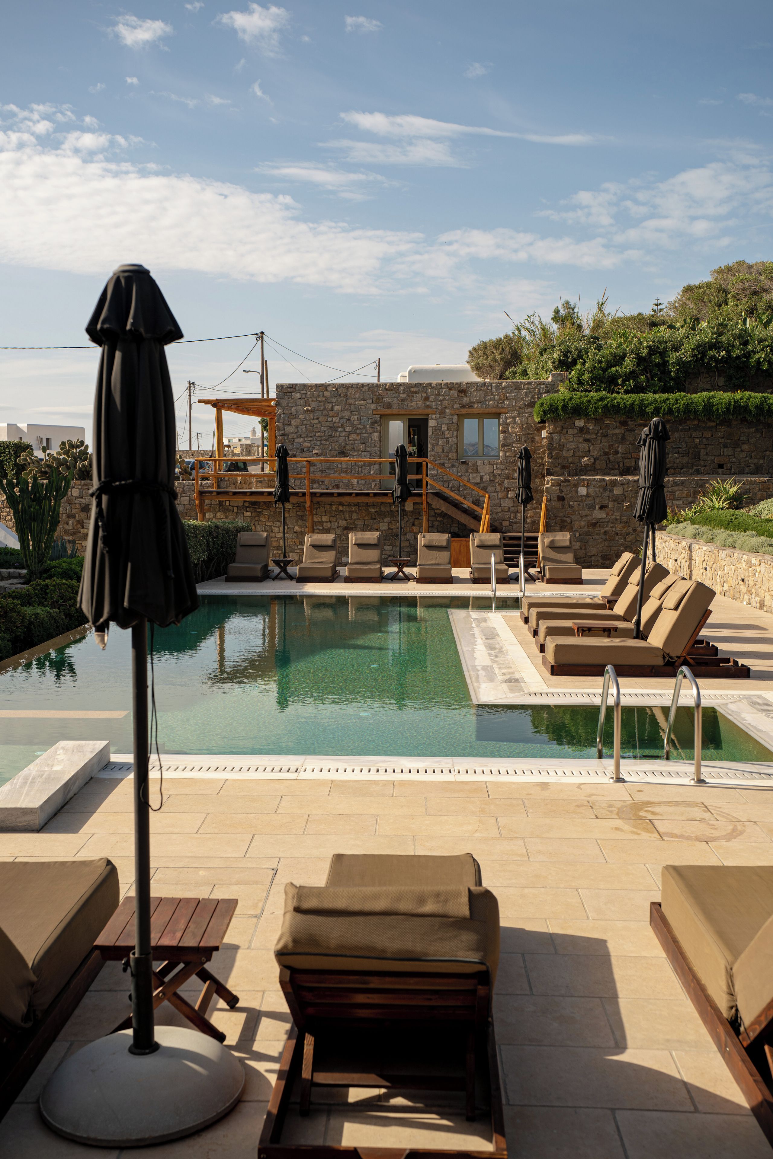 Zona de piscina con sillones de descanso y sombrillas, rodeada de muros de piedra y follaje.