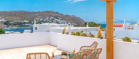 A terrace with wicker chairs and a table, overlooking a coastal town with white buildings and a view of the sea.