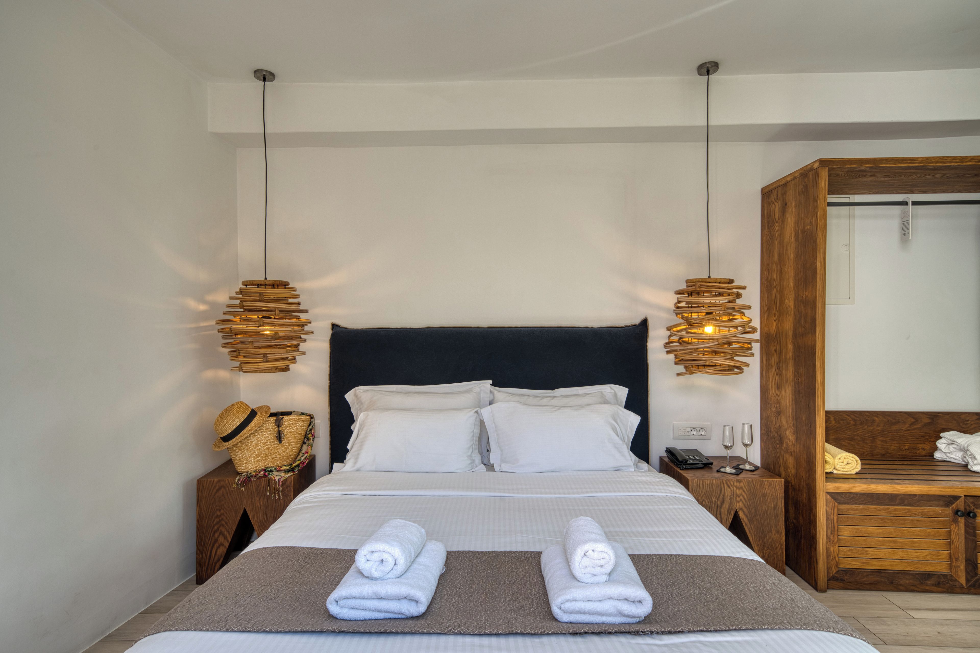 A neatly made bed with two rolled towels, a wooden nightstand with a lamp, and a wooden wardrobe.