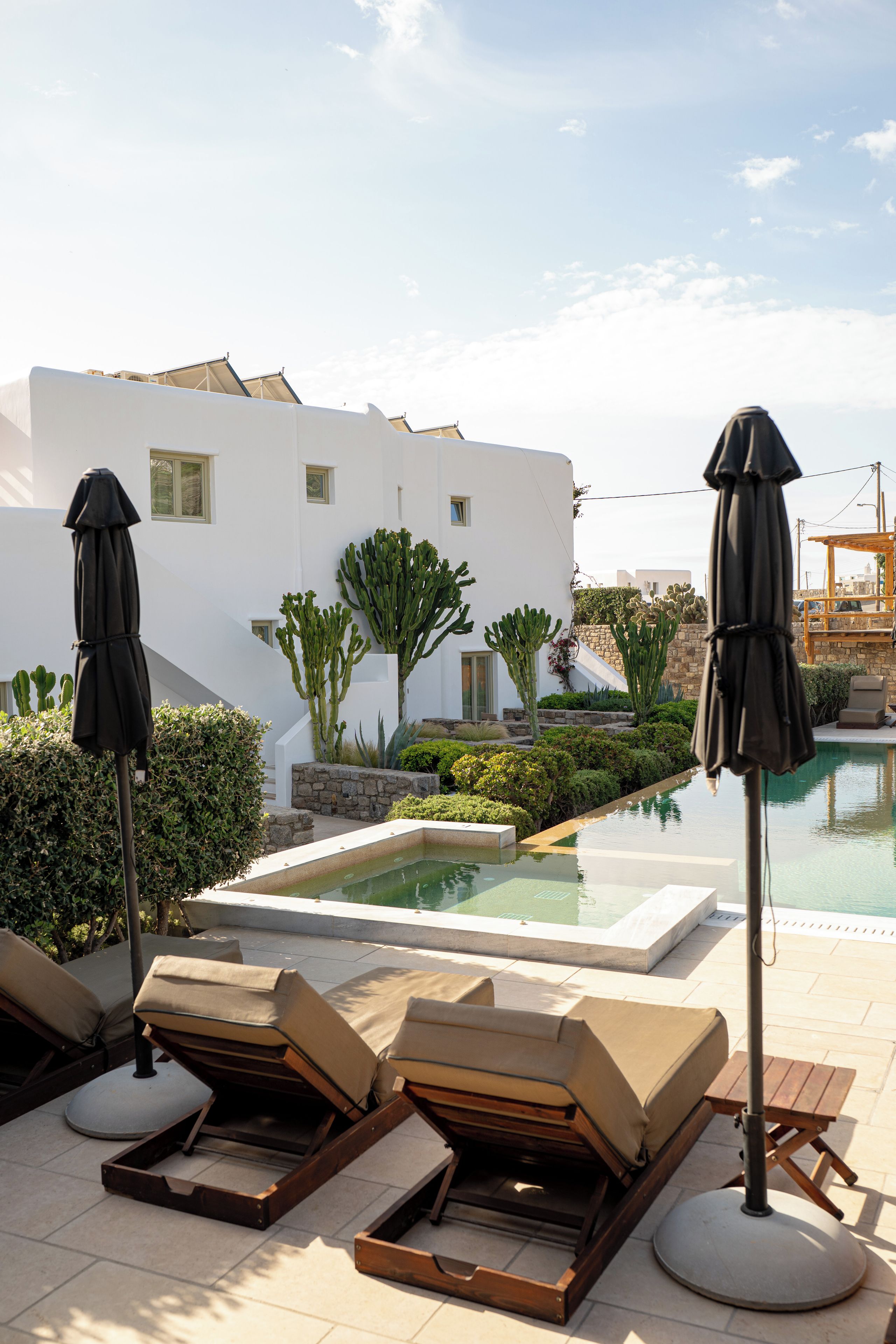 Zona de piscina con sillones de descanso y sombrillas, piscina y un edificio blanco con pared de piedra y cactus.