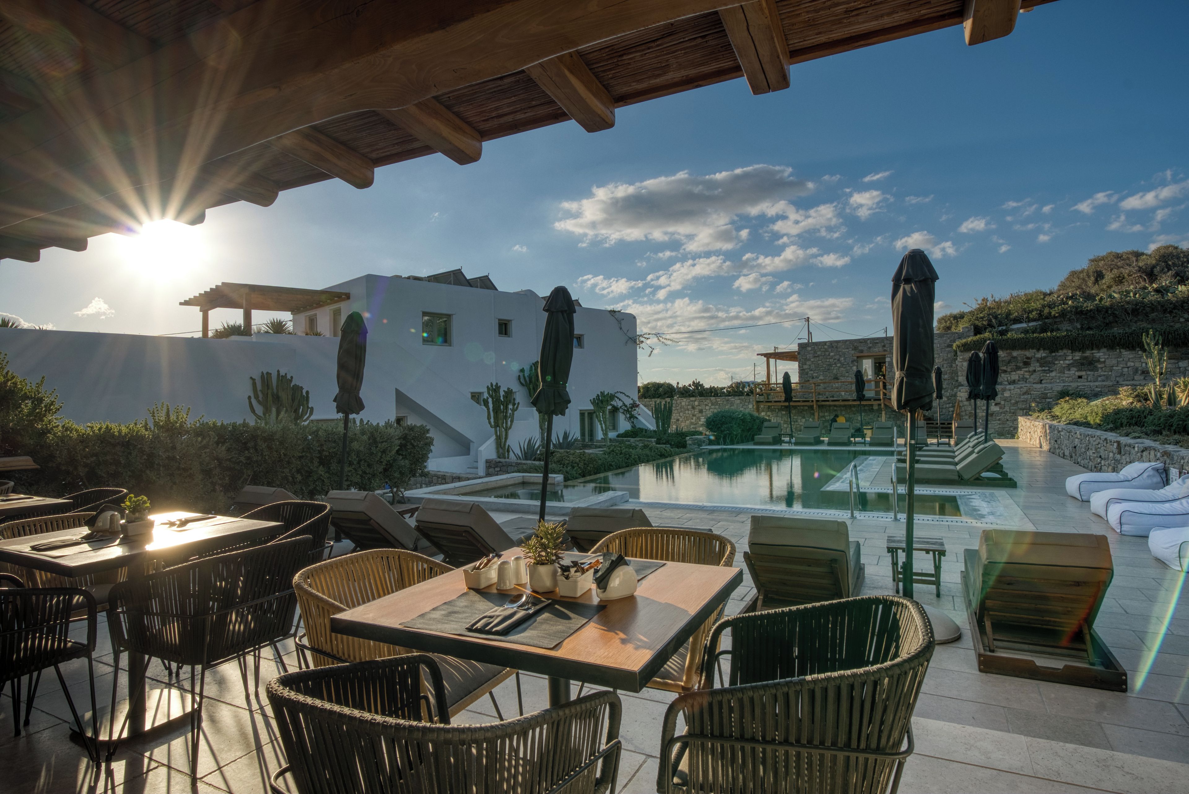 A sunlit outdoor dining area with a pool, white building, and stone wall in the background.