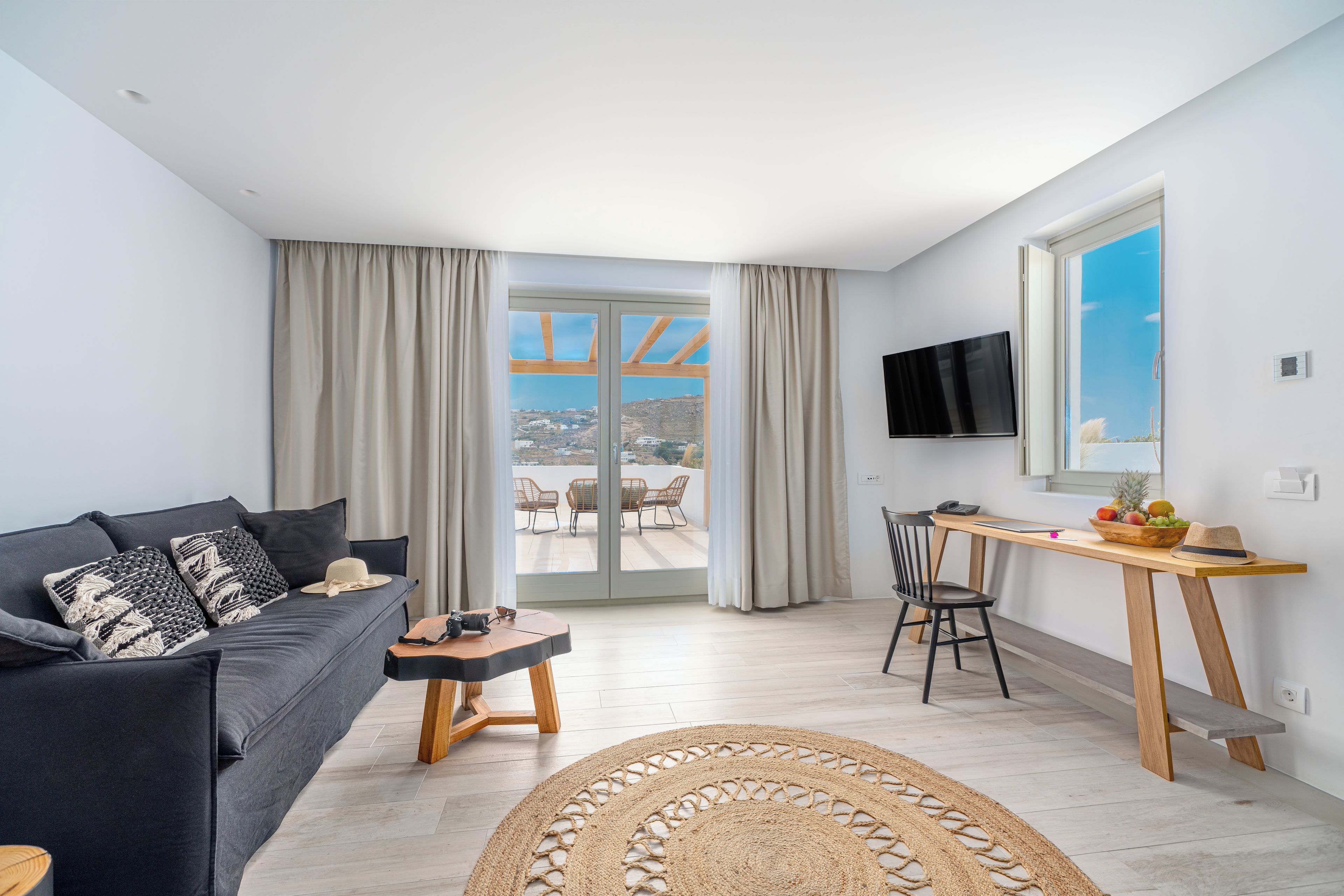 A modern living room with a sofa, a small coffee table, a dining area with a wooden table and chairs, a TV mounted on the wall, and a large window with curtains.