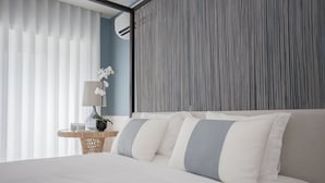 Ein ordentlich bezogenes Bett mit weißen und grauen Kissen, ein runder Nachttisch mit Lampe und Pflanze, und eine gestreifte Wand dahinter.