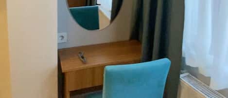 Una habitación pequeña con un escritorio de madera, una silla azul, un espejo redondo y un ventanal con cortinas.