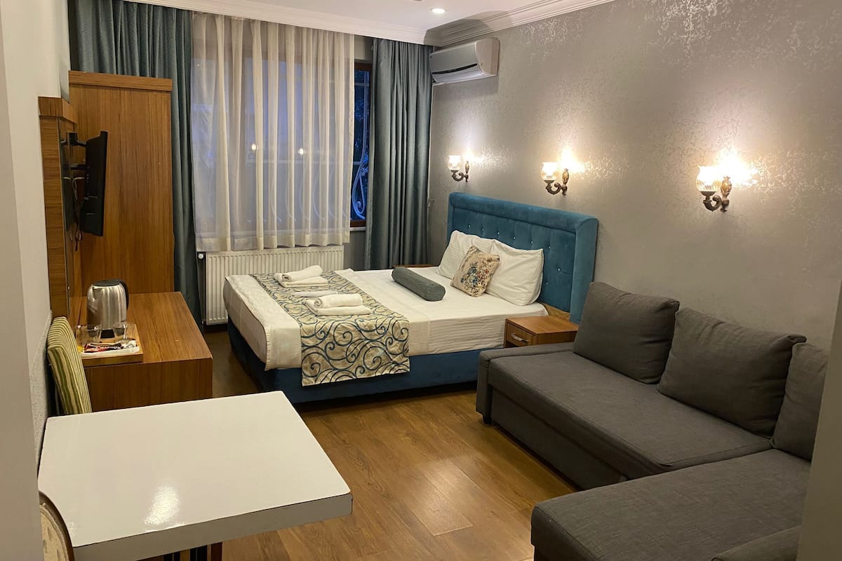 A hotel room with a bed, a sofa, a small table, an air conditioning unit, and a television.