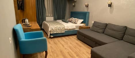 Habitación de hotel compacta con cama, sofá, sillón, una mesita con hervidor y un cubo de basura.