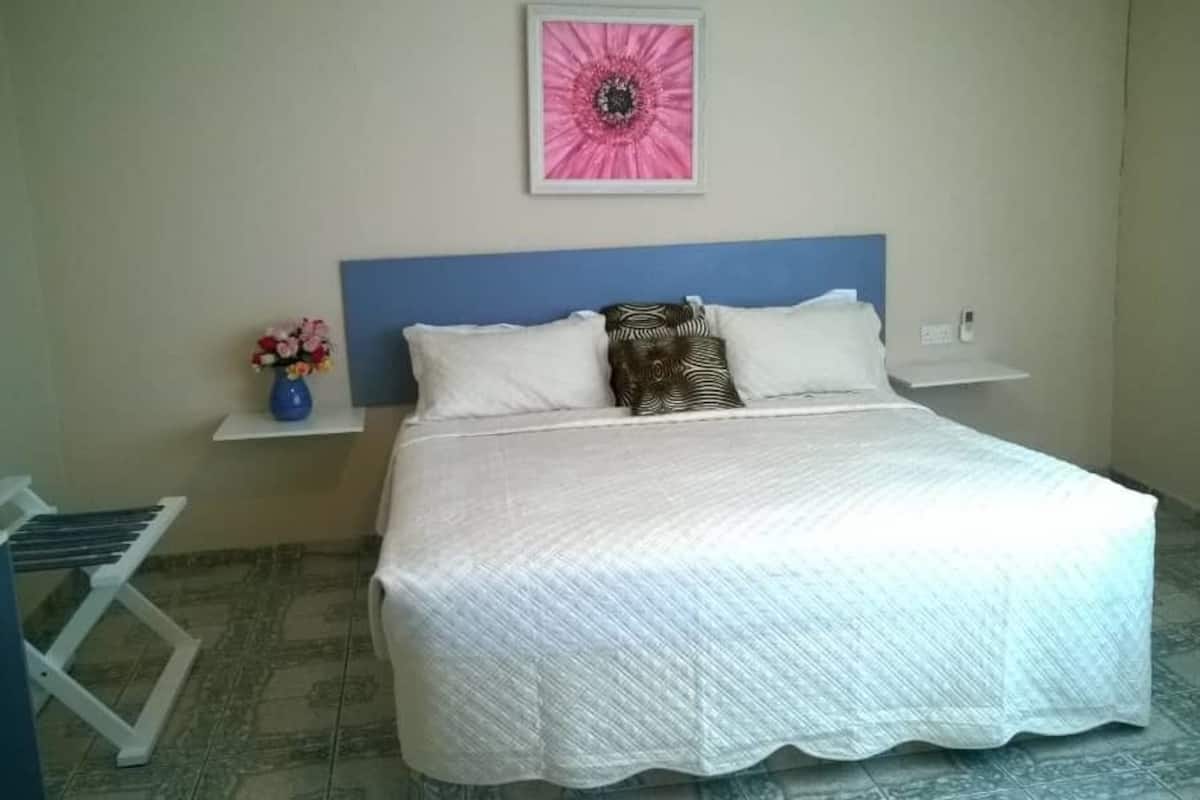 Un lit bien fait, recouvert d’une couvre-lit blanche et agrémenté d’oreillers, avec une tête de lit bleue, un tableau représentant une fleur rose accroché au mur, et une petite étagère sur laquelle est posé un vase de fleurs.