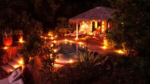 Estructura con techo de paja, con área de estar exterior y piscina, rodeada de plantas en macetas e iluminación ambiental.