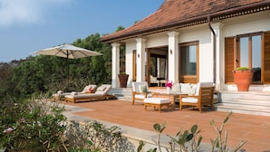 Una villa con patio de baldosas, mobiliario exterior y un gran parasol.