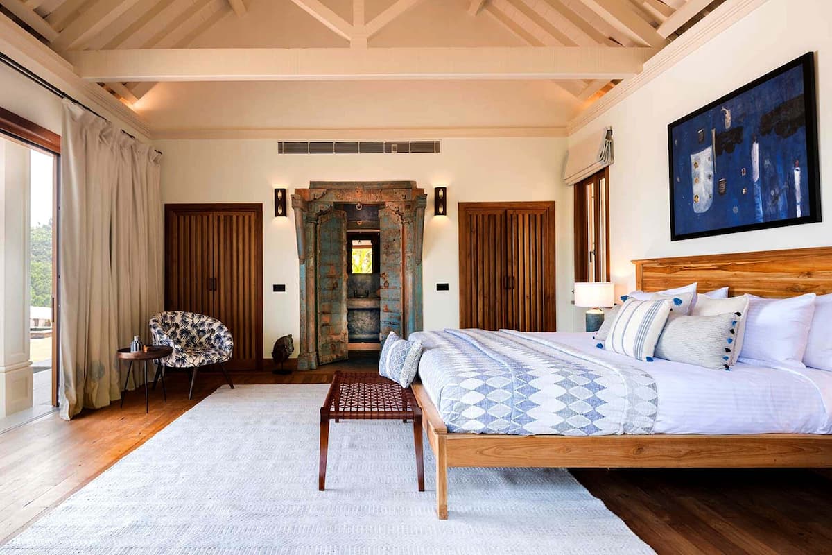 A spacious bedroom with a large bed, a wooden bench, a chair, and a view through a door.