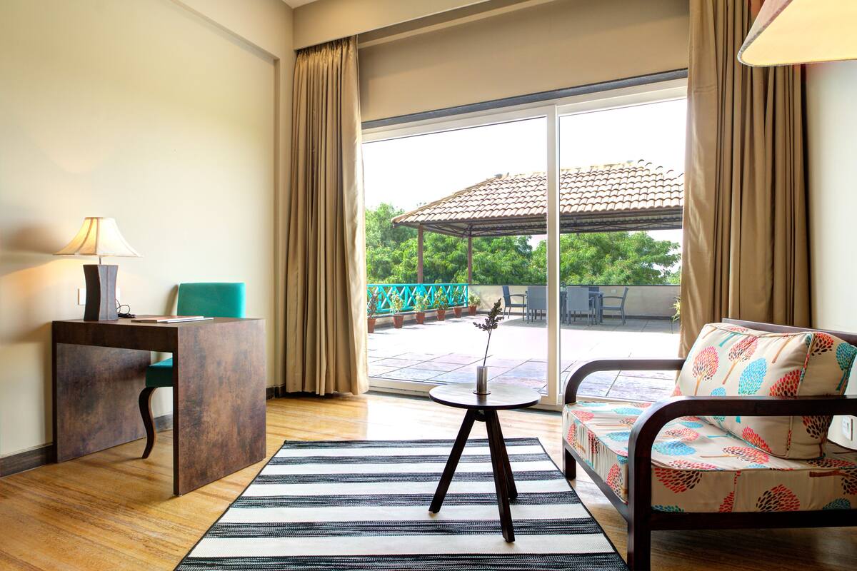 Habitación de hotel con escritorio, sofá, una pequeña mesa con un florero y vistas a un jardín y un edificio.