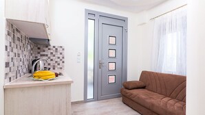 A compact living space with a kitchenette, a grey door with small windows, a brown sofa, and a window with sheer curtains. - Lena Apartments (Thasos)