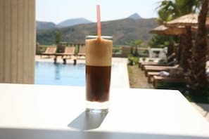 Un vaso de café helado con pajita, sobre una mesa junto a una piscina con sillones de descanso y una vista de la montaña al fondo.