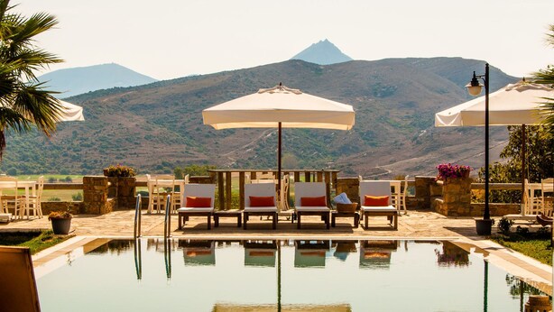 A pool with lounge chairs and umbrellas, surrounded by stone walls and a view of a mountain. - Anemos Hotel (Gökçeada)