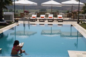 Zona de piscina con sillones de descanso y sombrillas, una parrilla y vistas al campo.