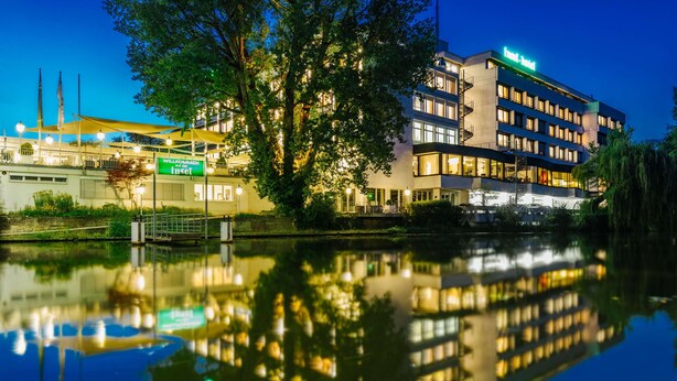 Ein Hotel mit Spiegelung im Wasser, das moderne Architektur, mehrere Stockwerke und ein sichtbares Firmenschild zeigt.