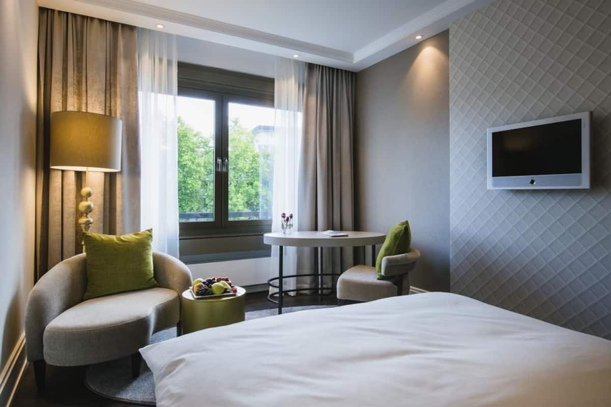 Quarto de hotel com cama, mesa redonda, duas cadeiras, televisão e janela com cortinas.