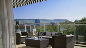 Ein Balkon mit Korbmöbeln, einem Glastisch und Blick auf eine Stadtlandschaft und Hügel.