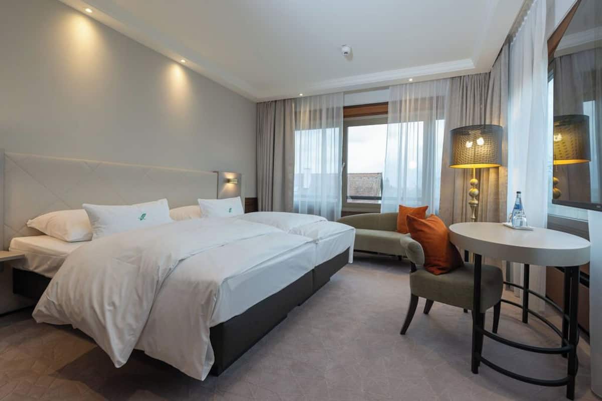 Quarto de hotel com uma cama grande, um sofá, uma mesa pequena e uma janela com cortinas.