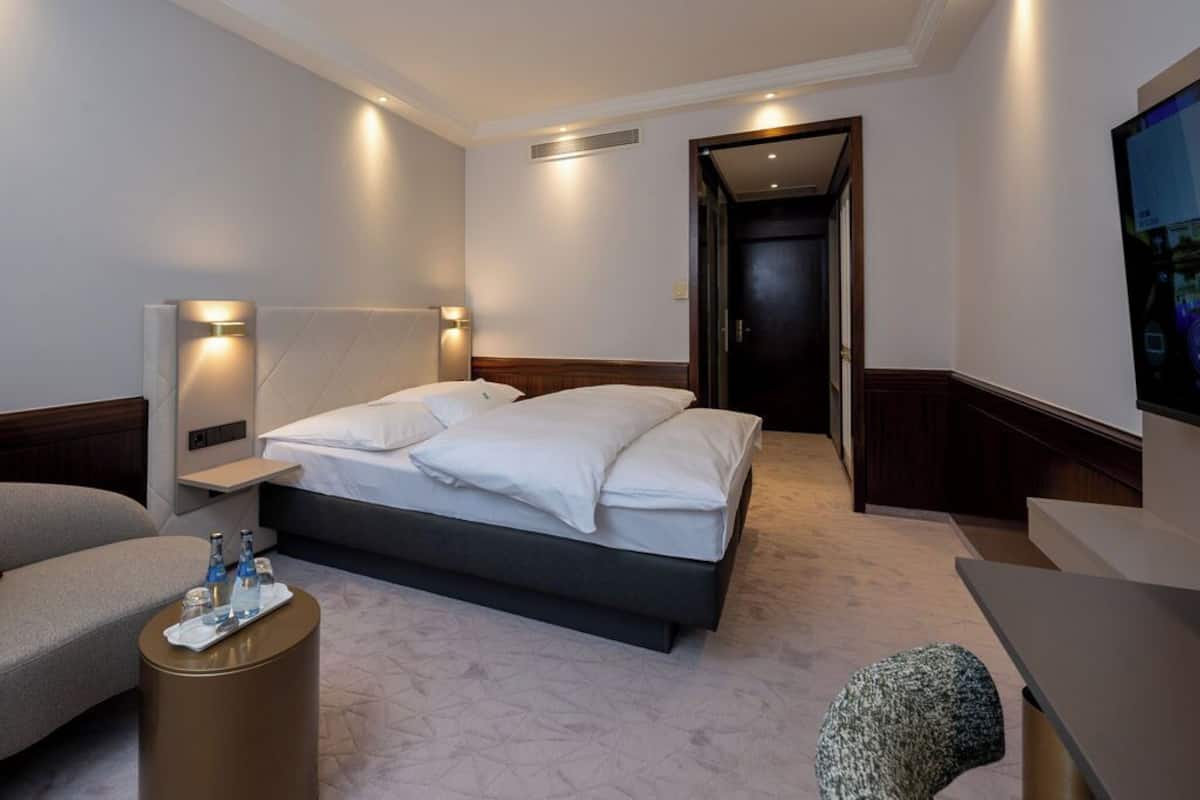Quarto de hotel moderno com uma cama grande, mesas de cabeceira, um espaço de estar e uma televisão.