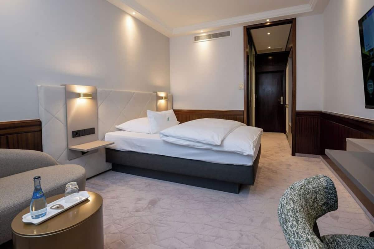 Quarto de hotel com cama, mesas de cabeceira, uma cadeira e uma pequena mesa com garrafa e copo.