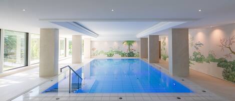 Una piscina interior moderna con agua cristalina y azul, rodeada de grandes ventanales y un mural de motivos naturales en las paredes.
