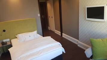 A hotel room with a bed, a bedside table with a phone, a TV mounted on the wall, and a door leading to another room.
