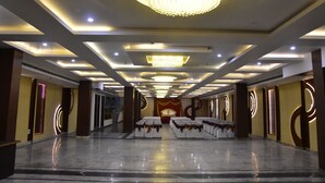 A spacious hall with a chandelier, recessed lighting, and rows of tables with white tablecloths and brown centerpieces. - Ishanika Hotel (Lucknow)
