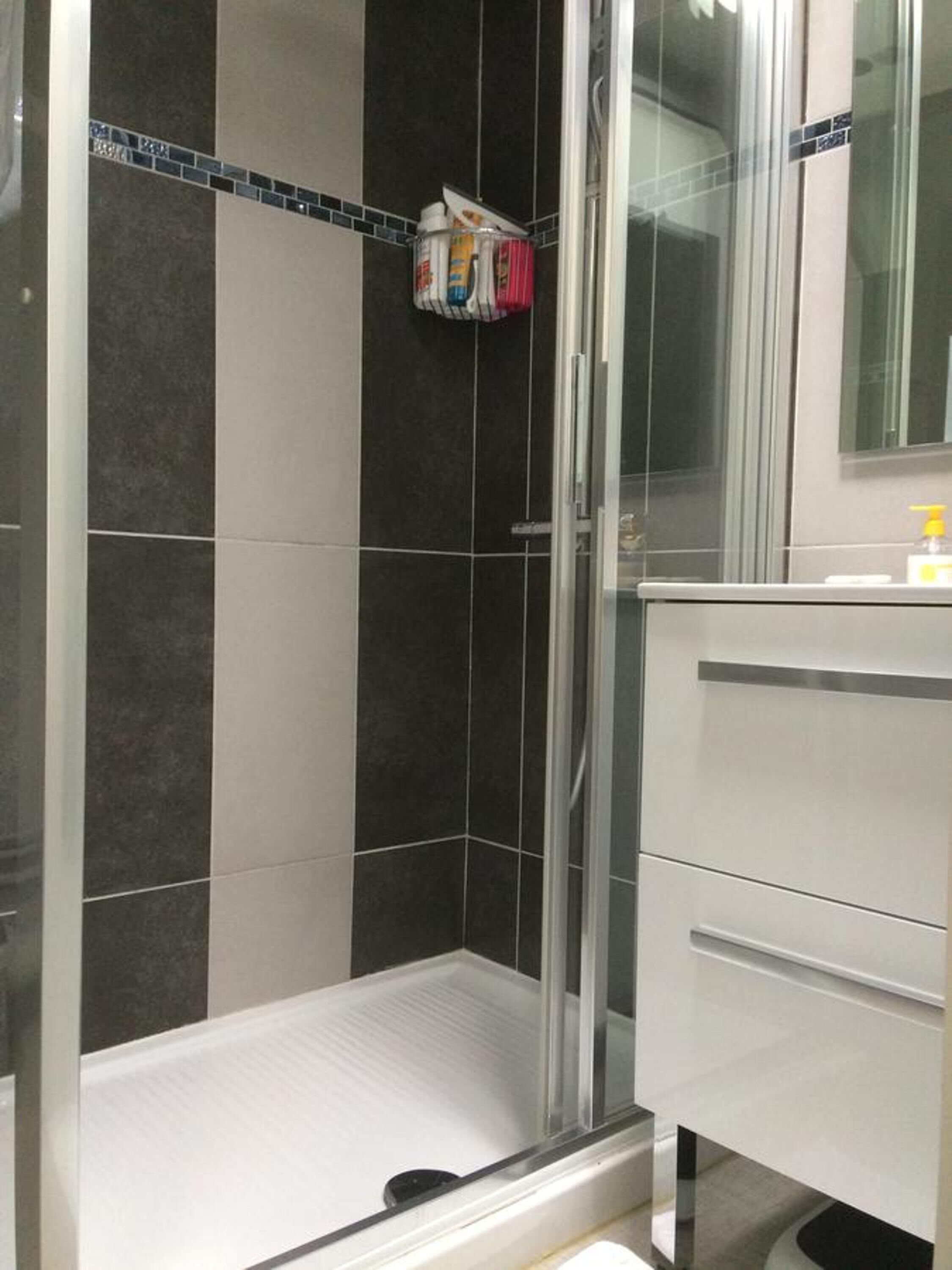 Une salle de bain moderne avec une douche à l’italienne en verre, des murs carrelés aux tons foncés et clairs, et un meuble-lavabo blanc avec un miroir.
