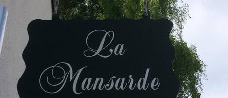A black sign with the text "La Mansarde" in white cursive font, hanging from a decorative metal frame.