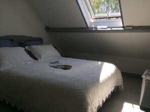 Une chambre avec un puits de lumière, un lit recouvert d’une couverture blanche tricotée et une fenêtre donnant sur des arbres.