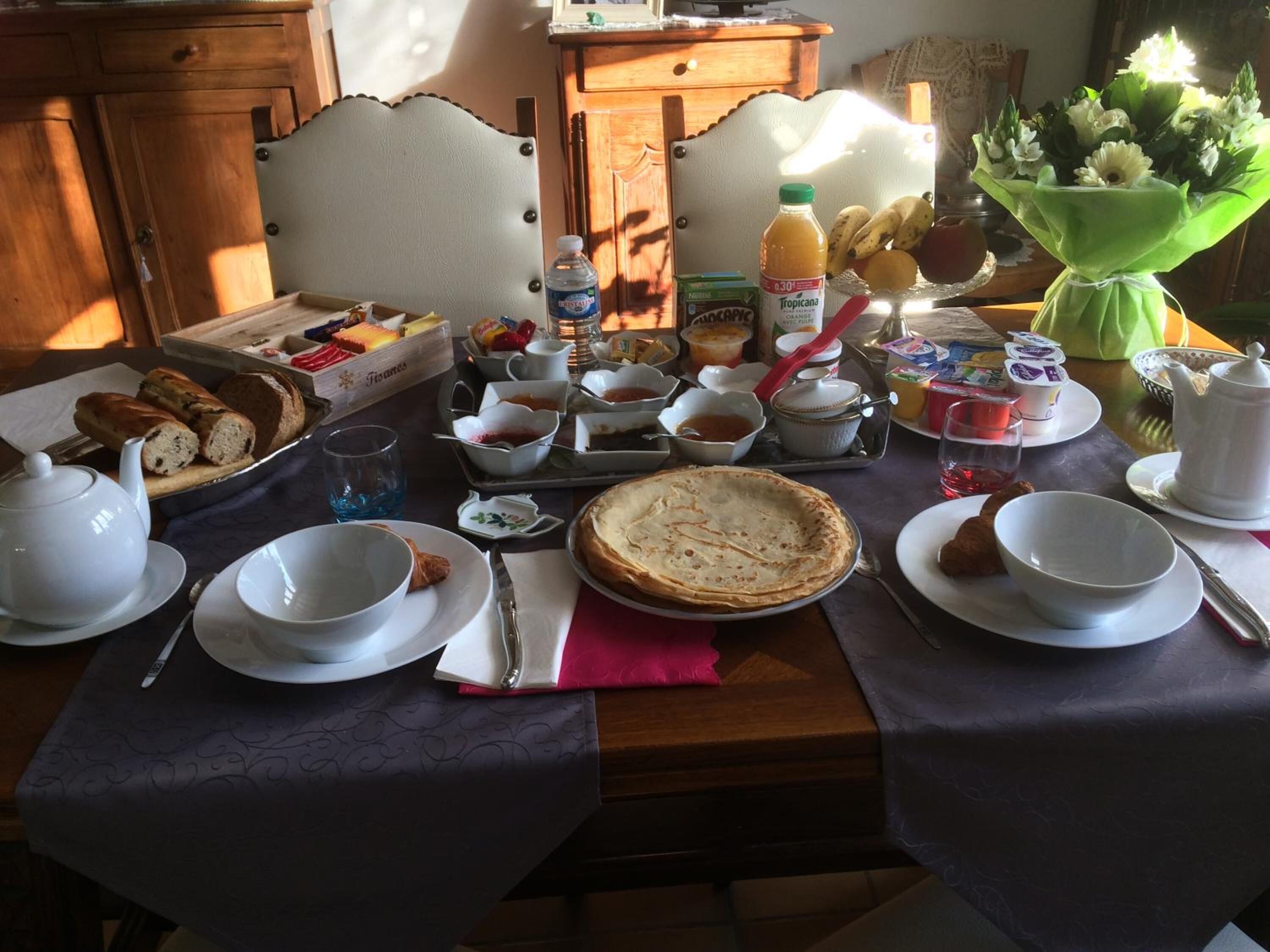 Une table de petit-déjeuner avec divers aliments, notamment du pain, des viennoiseries et une tarte, ainsi que plusieurs tasses et une bouteille de jus de fruits.