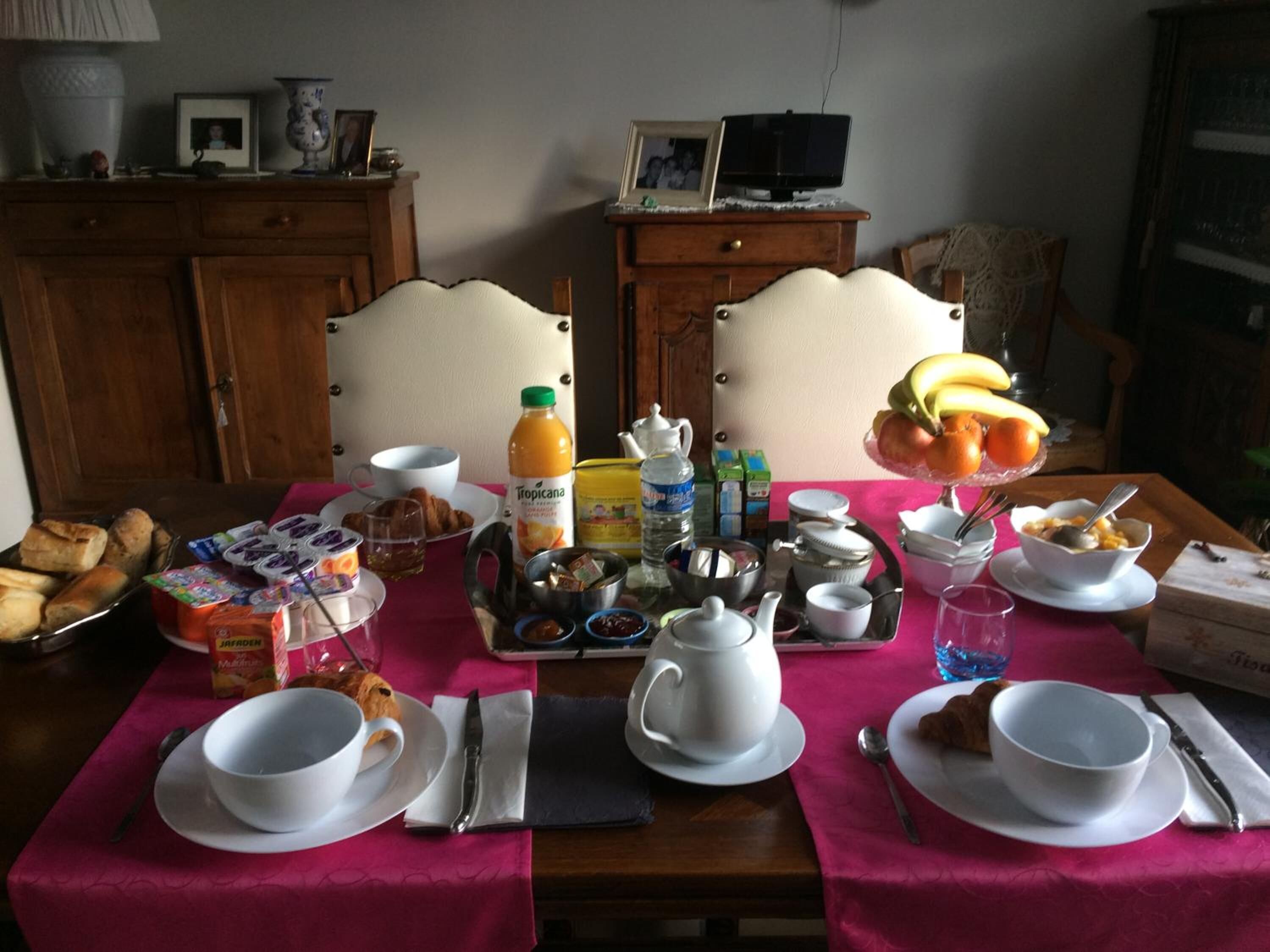 Mesa de desayuno servida con pan, cruasanes y fruta, con mantel rosa.
