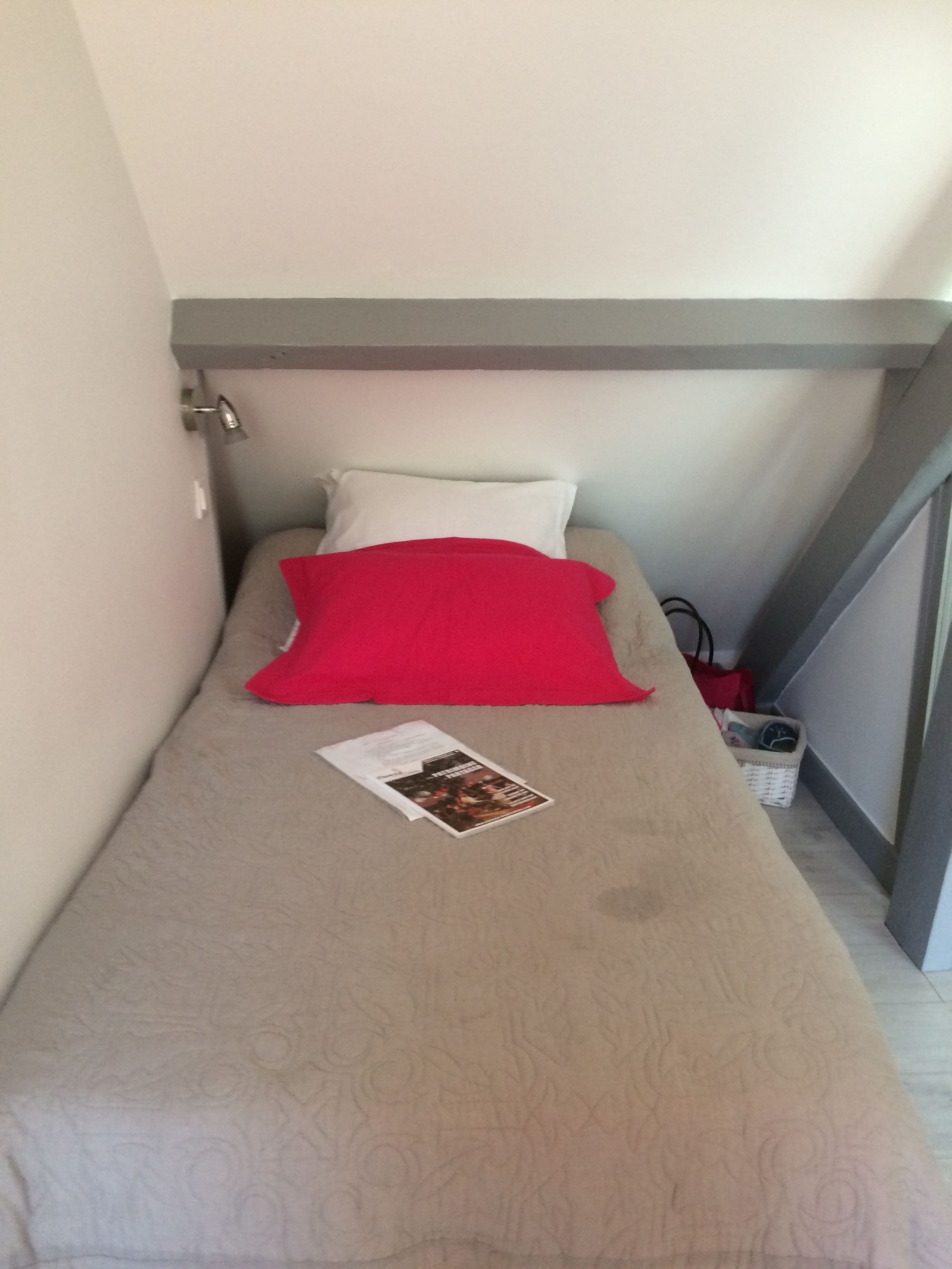 Una habitación pequeña y estrecha con una cama individual, un almohadón rojo y una revista sobre la cama.