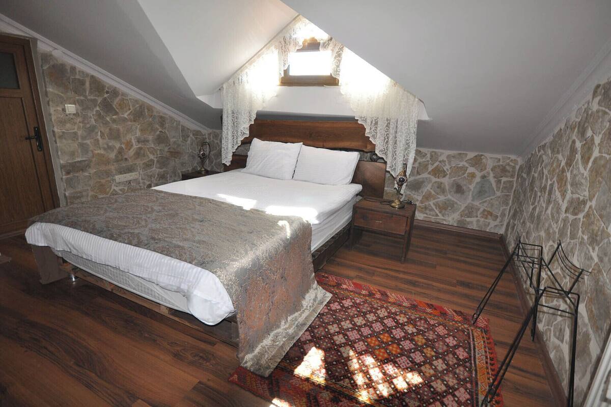 A bedroom with a stone wall, wooden floor, a bed with white bedding, a nightstand, and a patterned rug.