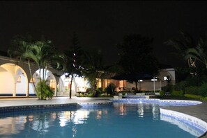 Zona de piscina nocturna con borde de azulejos, palmeras y un edificio con arcos al fondo.