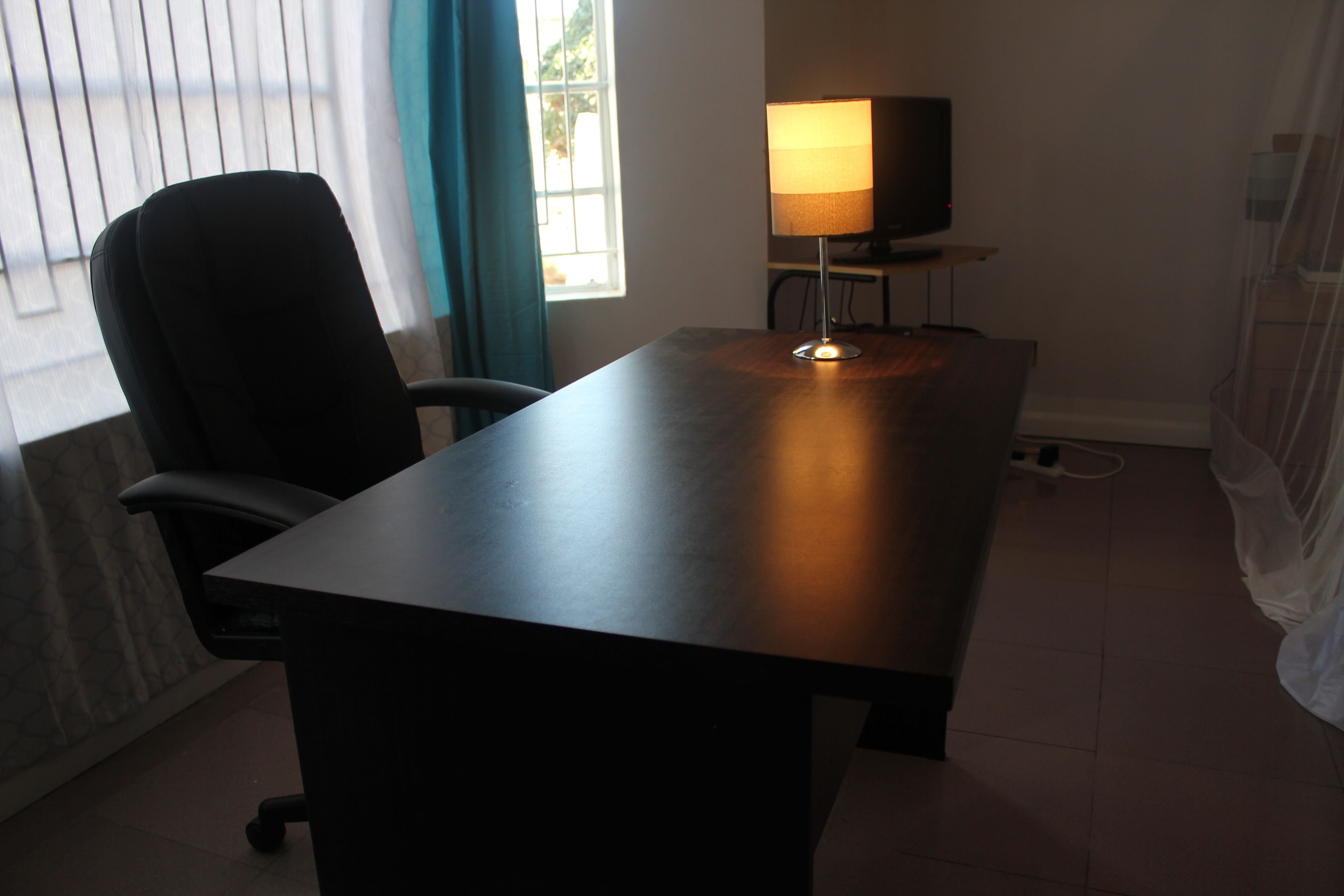 Una habitación con escritorio, silla, lámpara, televisor y ventana con cortinas.
