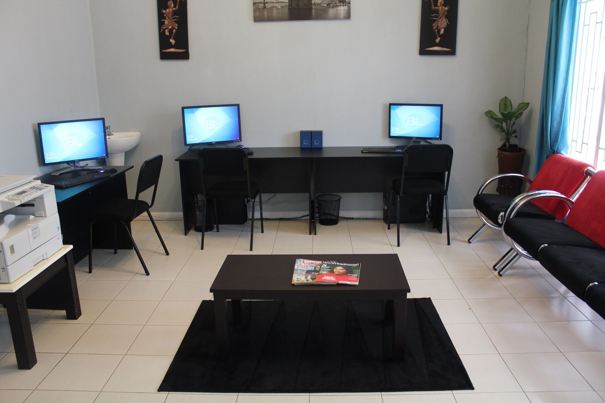 A modern office space with two desks, each with a computer monitor, black chairs, a black coffee table with a magazine, and a red sofa.