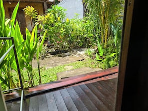 A wooden deck with a view of a garden with various plants and a brick pathway. - Timbis Homestay Bali (Kutuh)