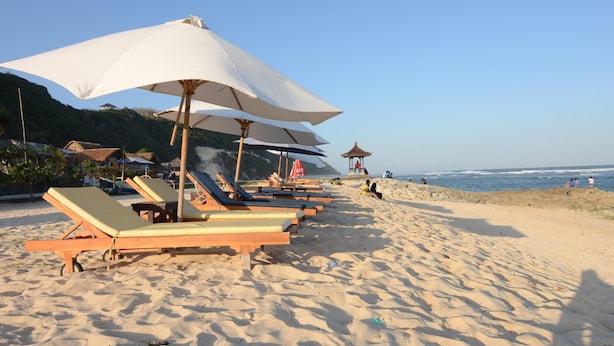 A beach with sun loungers and umbrellas, a rocky outcrop, and the ocean in the background. - Timbis Homestay Bali (Kutuh)