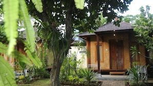 A small, single-story building with a tiled roof and a wooden door. It is surrounded by lush greenery, including various plants and trees. - Timbis Homestay Bali (Kutuh)