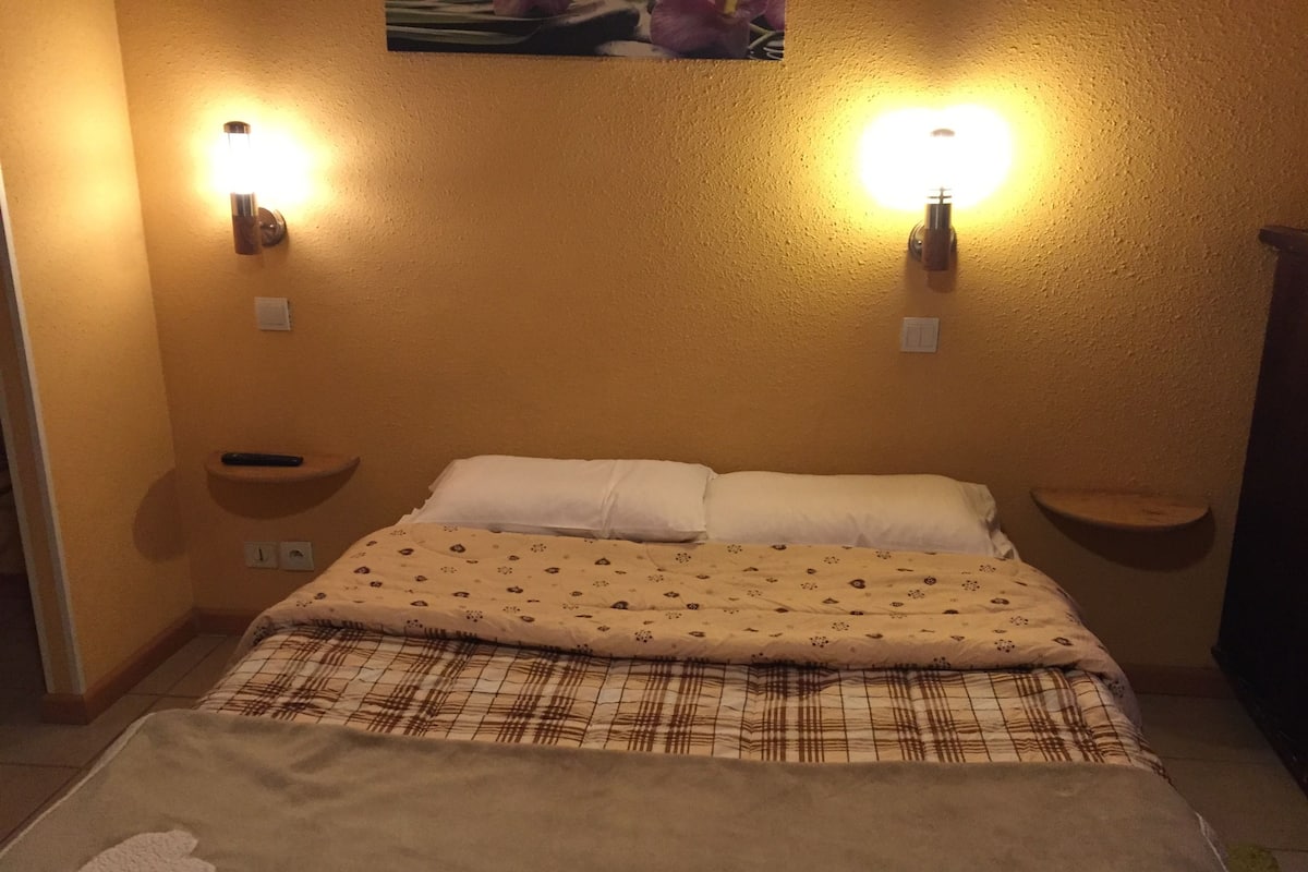 Un dormitori amb un llit, dues làmpades murals, una petita estant i un rellotge de paret.